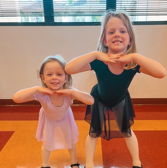 Two young ballet students pose with hands under their chins, one in pink and one in black, in a dance studio
